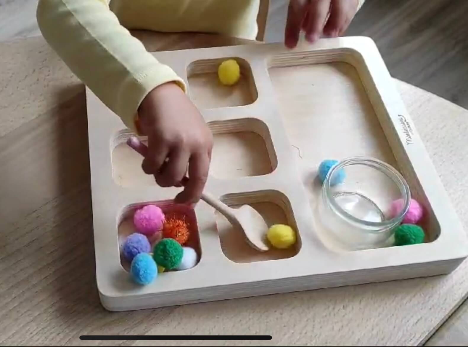 Montessori - Activité - GDL La main d'un enfant qui s'entraine au transvasement avec une cuillère et des pompons colorés